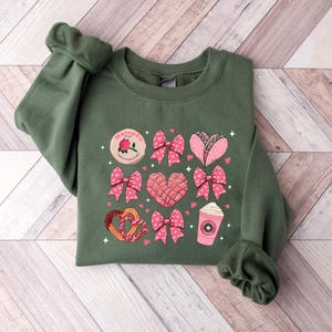 May include: A green sweatshirt with a pink and white design featuring hearts, bows, and various Mexican treats. The text "Ma&ntilde;anitas" is printed on the sweatshirt.