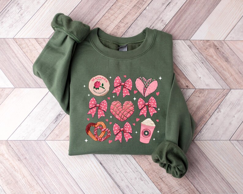 May include: A green sweatshirt with a pink and white design featuring hearts, bows, and various Mexican treats. The text "Ma&ntilde;anitas" is printed on the sweatshirt.