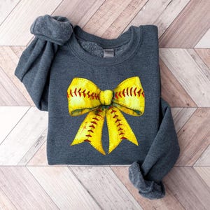 May include: A dark gray sweatshirt with a yellow and red softball bow design. The bow is made to look like a softball with red stitching.