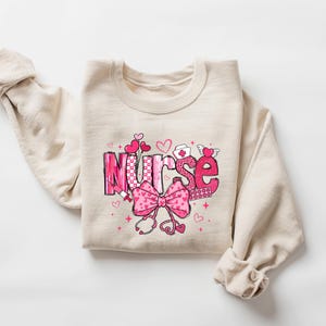 May include: A beige sweatshirt with a pink and white graphic design that says "Nurse" with a pink bow and other medical symbols.