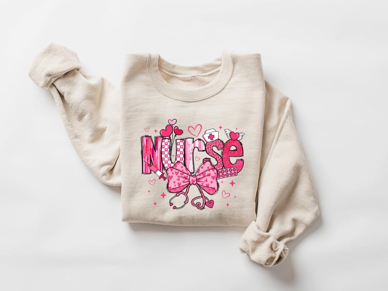 May include: A beige sweatshirt with a pink and white graphic design that says "Nurse" with a pink bow and other medical symbols.