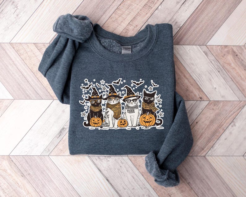 May include: A dark gray sweatshirt with a graphic of four cats wearing witch hats and holding pumpkins. The cats are wearing glasses and have different colored fur. The graphic is surrounded by bats and snowflakes.