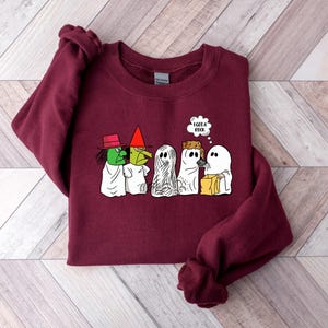 Puede incluir: Una sudadera color burdeos con un estampado gr&aacute;fico de cuatro fantasmas y una bruja. Los fantasmas llevan disfraces y llevan bolsas de golosinas. La bruja lleva un sombrero y sostiene una escoba. El texto "Tengo un libro" est&aacute; en un globo de pensamiento sobre los fantasmas.