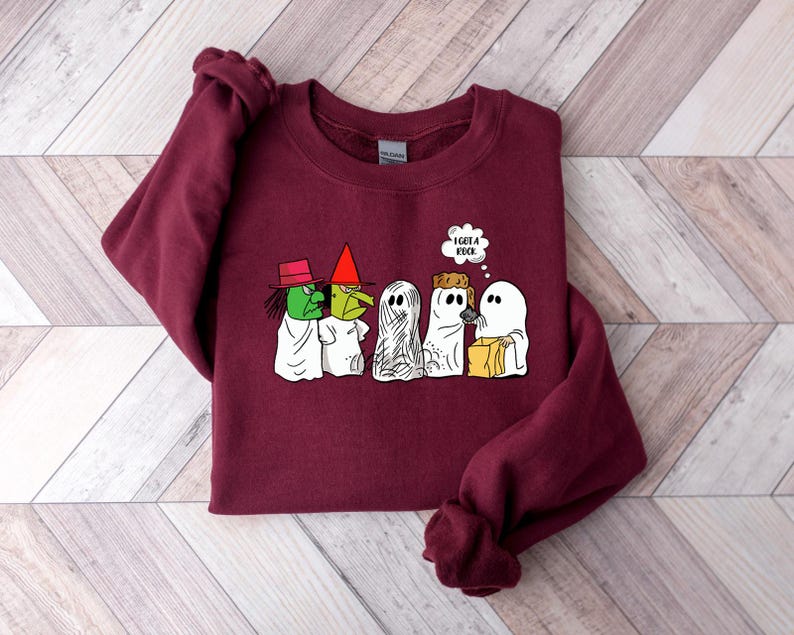 Puede incluir: Una sudadera color burdeos con un estampado gr&aacute;fico de cuatro fantasmas y una bruja. Los fantasmas llevan disfraces y llevan bolsas de golosinas. La bruja lleva un sombrero y sostiene una escoba. El texto "Tengo un libro" est&aacute; en un globo de pensamiento sobre los fantasmas.