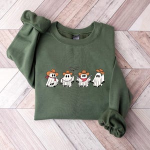 May include: An olive green sweatshirt featuring a row of four cartoon ghosts dressed as cowboys. Each ghost wears a brown cowboy hat and a red bandana. The ghosts are white with black eyes and a black mouth.