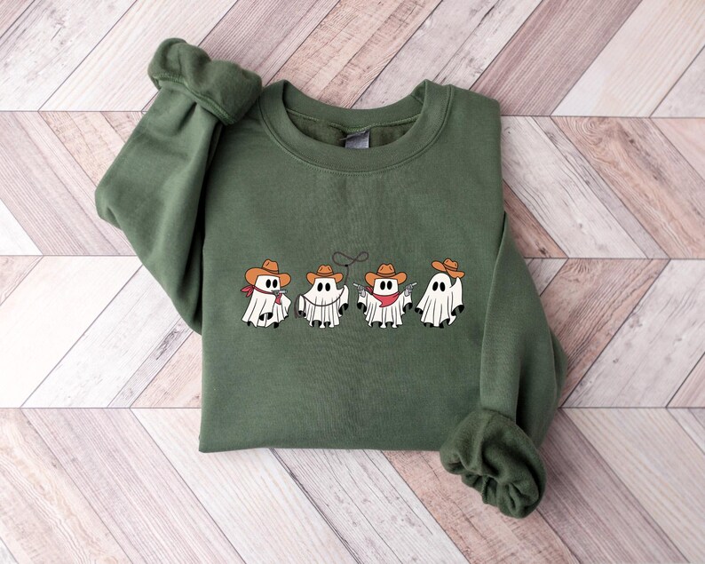 May include: An olive green sweatshirt featuring a row of four cartoon ghosts dressed as cowboys. Each ghost wears a brown cowboy hat and a red bandana. The ghosts are white with black eyes and a black mouth.
