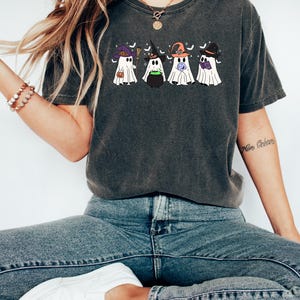 Puede incluir: Una camiseta gris oscuro con un gr&aacute;fico de cuatro fantasmas vestidos de brujas. Los fantasmas llevan sombreros de bruja y sostienen objetos con tem&aacute;tica de Halloween como un caldero, una escoba, una calabaza y un libro. La camiseta tiene un aspecto desgastado.