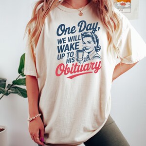 Puede incluir: Camiseta color crema con el texto "One Day, We Will Wake Up To His Obituary" en azul marino y rojo. La camiseta presenta una ilustraci&oacute;n vintage de una mujer sosteniendo una taza de caf&eacute;. Una prenda casual para el d&iacute;a a d&iacute;a.