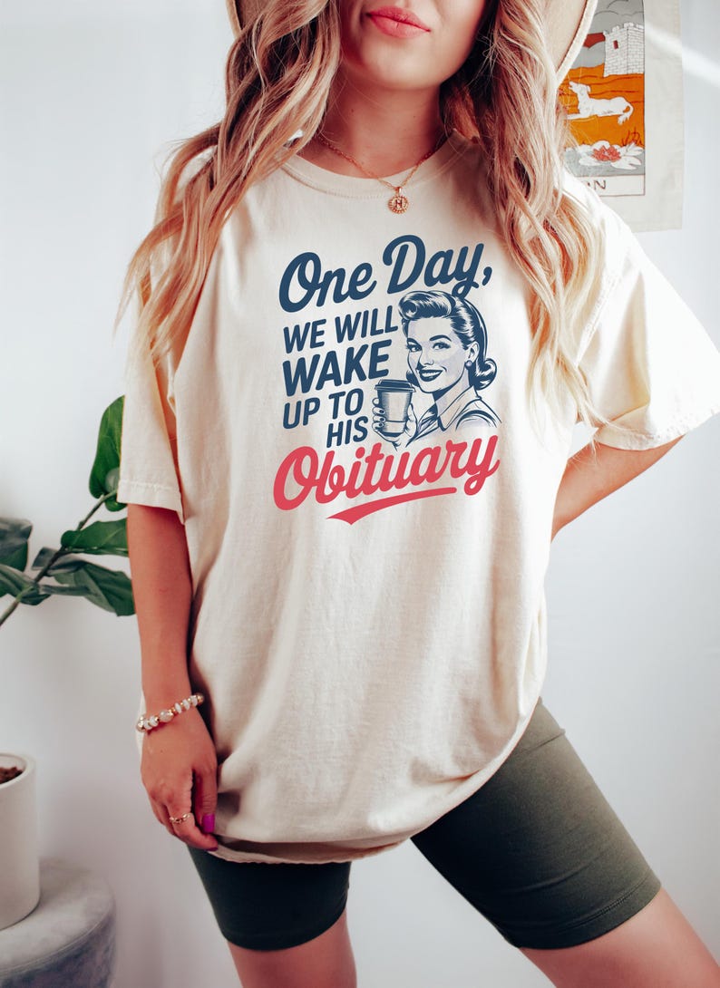 Puede incluir: Camiseta color crema con el texto "One Day, We Will Wake Up To His Obituary" en azul marino y rojo. La camiseta presenta una ilustraci&oacute;n vintage de una mujer sosteniendo una taza de caf&eacute;. Una prenda casual para el d&iacute;a a d&iacute;a.