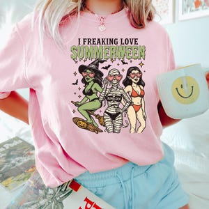 May include: Pink t-shirt with the text "I Freaking Love Summerween" and a graphic of three cartoon women dressed as a witch, mummy, and bikini-clad woman. The witch is riding a skateboard with a pumpkin. A mug with a smiley face is held in the hand.