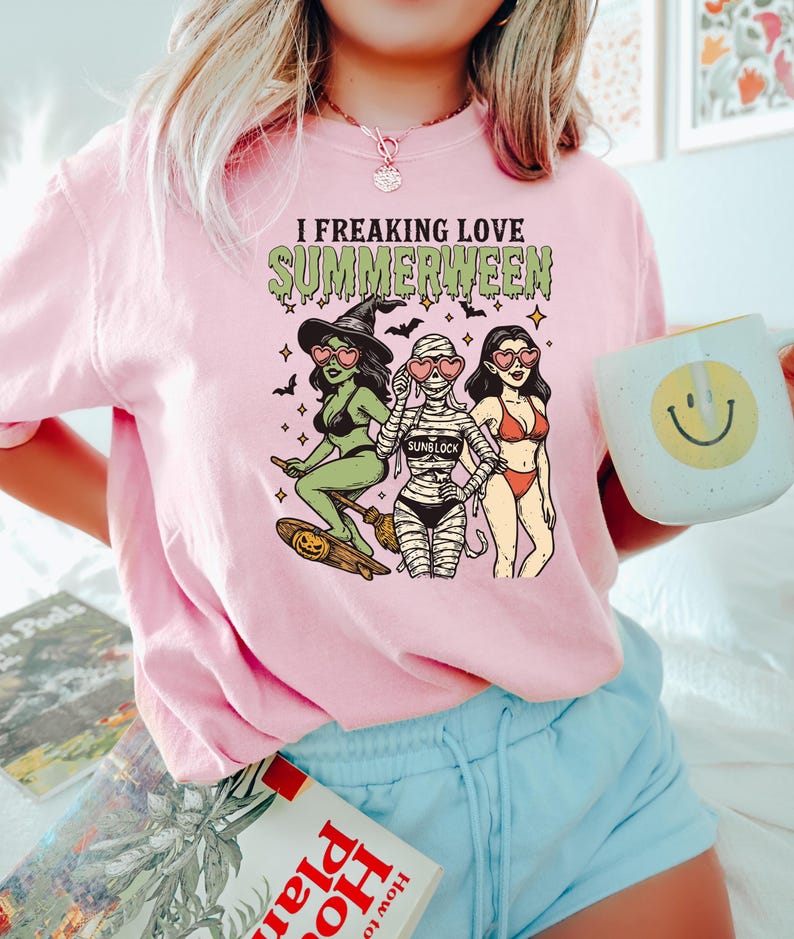 May include: Pink t-shirt with the text "I Freaking Love Summerween" and a graphic of three cartoon women dressed as a witch, mummy, and bikini-clad woman. The witch is riding a skateboard with a pumpkin. A mug with a smiley face is held in the hand.