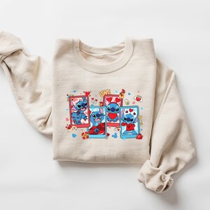 May include: A beige sweatshirt with a graphic print of Stitch from Lilo & Stitch. Stitch is shown in four different poses, each with a red heart and surrounded by hearts and other Valentine's Day themed graphics. The text "LOVE" is printed above the graphic.