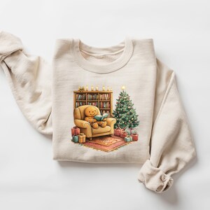 May include: A beige sweatshirt with a Christmas-themed graphic. The graphic features a gingerbread person sitting in a chair, reading a book, next to a Christmas tree and a bookshelf.