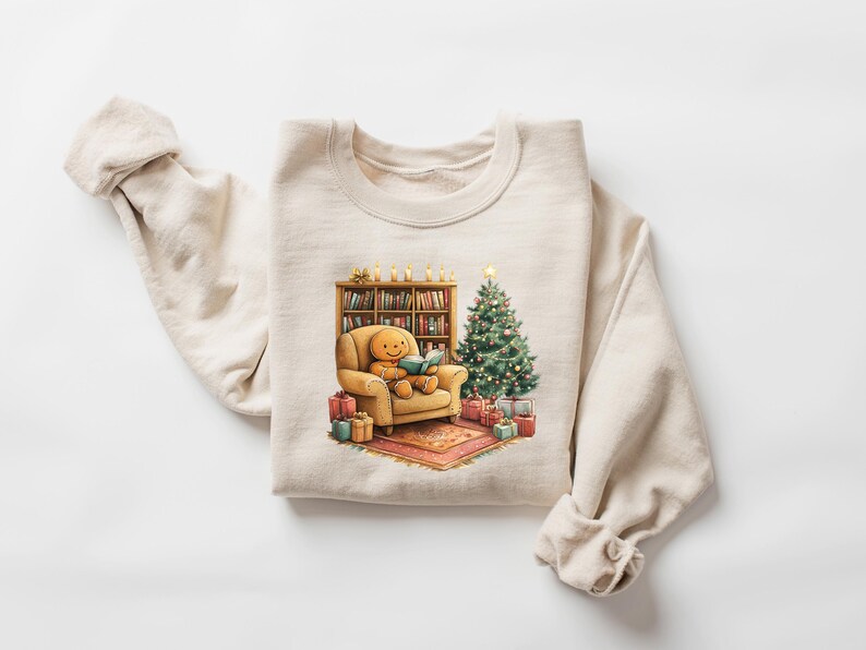 May include: A beige sweatshirt with a Christmas-themed graphic. The graphic features a gingerbread person sitting in a chair, reading a book, next to a Christmas tree and a bookshelf.