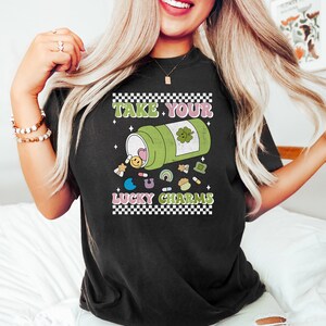 May include: Black t-shirt with a graphic of a green pill bottle with a four-leaf clover on it. The bottle is spilling out lucky charms, including a rainbow, a horseshoe, a shamrock, and a smiley face. The text "Take Your Lucky Charms" is printed on the shirt.