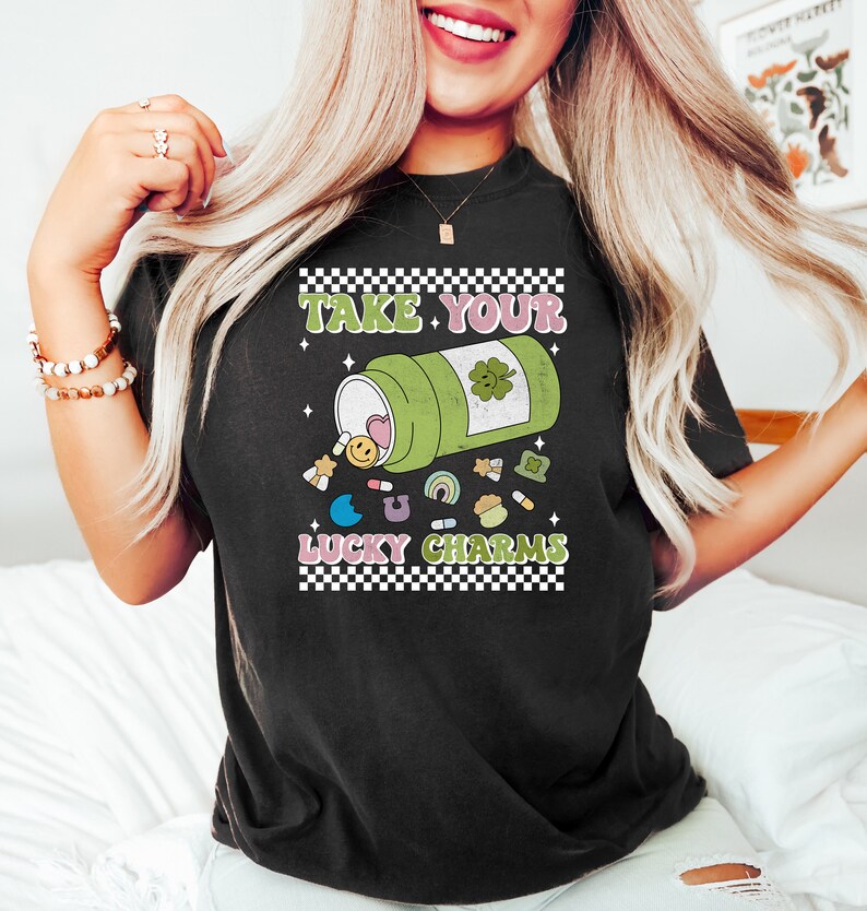 May include: Black t-shirt with a graphic of a green pill bottle with a four-leaf clover on it. The bottle is spilling out lucky charms, including a rainbow, a horseshoe, a shamrock, and a smiley face. The text "Take Your Lucky Charms" is printed on the shirt.