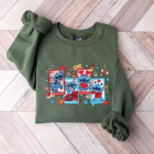 May include: Green sweatshirt with a graphic print of Stitch from Lilo & Stitch. The print features Stitch in four different poses with hearts and the word "LOVE".