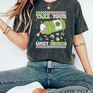 May include: A dark gray t-shirt with a graphic of a green pill bottle with the text "Take Your Lucky Charms" printed on it. The pill bottle is spilling out colorful charms, including a heart, a smiley face, a rainbow, a clover, and a horseshoe.