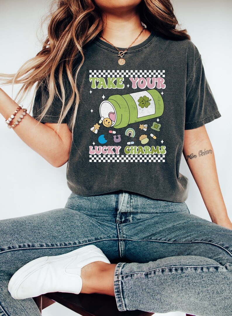 May include: A dark gray t-shirt with a graphic of a green pill bottle with the text "Take Your Lucky Charms" printed on it. The pill bottle is spilling out colorful charms, including a heart, a smiley face, a rainbow, a clover, and a horseshoe.