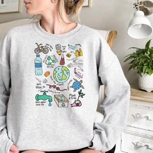 May include: A gray sweatshirt with a colorful graphic print of various environmental messages and illustrations. The graphic includes a bicycle, a water bottle, a turtle, a tree, and the words "Save the Planet", "Recycle", "Reduce", "Reuse", "Don't Litter", "Save Water Save Life", and "Plant a Tree".