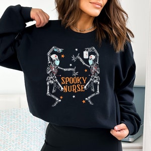 Puede incluir: Sudadera negra con un gr&aacute;fico de dos esqueletos vestidos de enfermeras con mascarillas. El texto "Spooky Nurse" est&aacute; impreso en naranja en la sudadera.