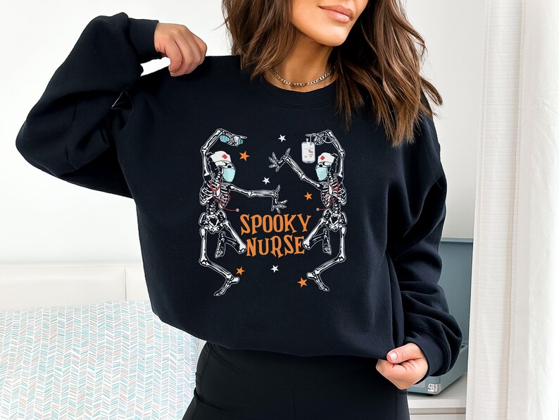 Puede incluir: Sudadera negra con un gr&aacute;fico de dos esqueletos vestidos de enfermeras con mascarillas. El texto "Spooky Nurse" est&aacute; impreso en naranja en la sudadera.