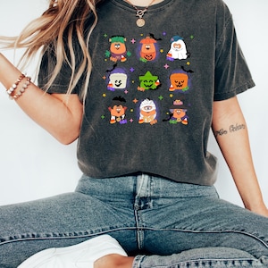 May include: A dark gray t-shirt with a vintage look featuring a Halloween-themed design of various cartoon characters dressed in costumes. The characters are holding candy buckets and wearing hats and accessories. The design includes a pumpkin, a ghost, a mummy, a witch, and a Frankenstein.