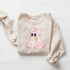 May include: Beige crewneck sweatshirt featuring a floral ghost graphic with a pink bow. The ghost design incorporates a patchwork of floral patterns in pink, yellow, and green. The sweatshirt has long sleeves and a relaxed fit, perfect for casual wear.
