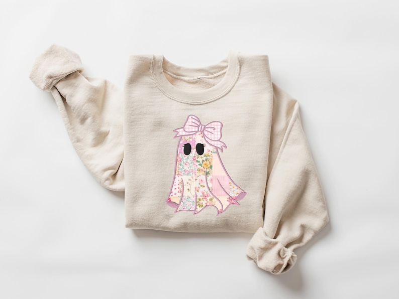 May include: Beige crewneck sweatshirt featuring a floral ghost graphic with a pink bow. The ghost design incorporates a patchwork of floral patterns in pink, yellow, and green. The sweatshirt has long sleeves and a relaxed fit, perfect for casual wear.