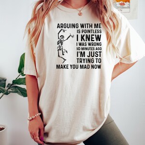 Puede incluir: Camiseta color crema con el texto "ARGUING WITH ME IS POINTLESS I KNEW I WAS WRONG 10 MINUTES AGO I'M JUST TRYING TO MAKE YOU MAD NOW" y un gr&aacute;fico de esqueleto. La camiseta se usa con pantalones cortos verde oscuro.