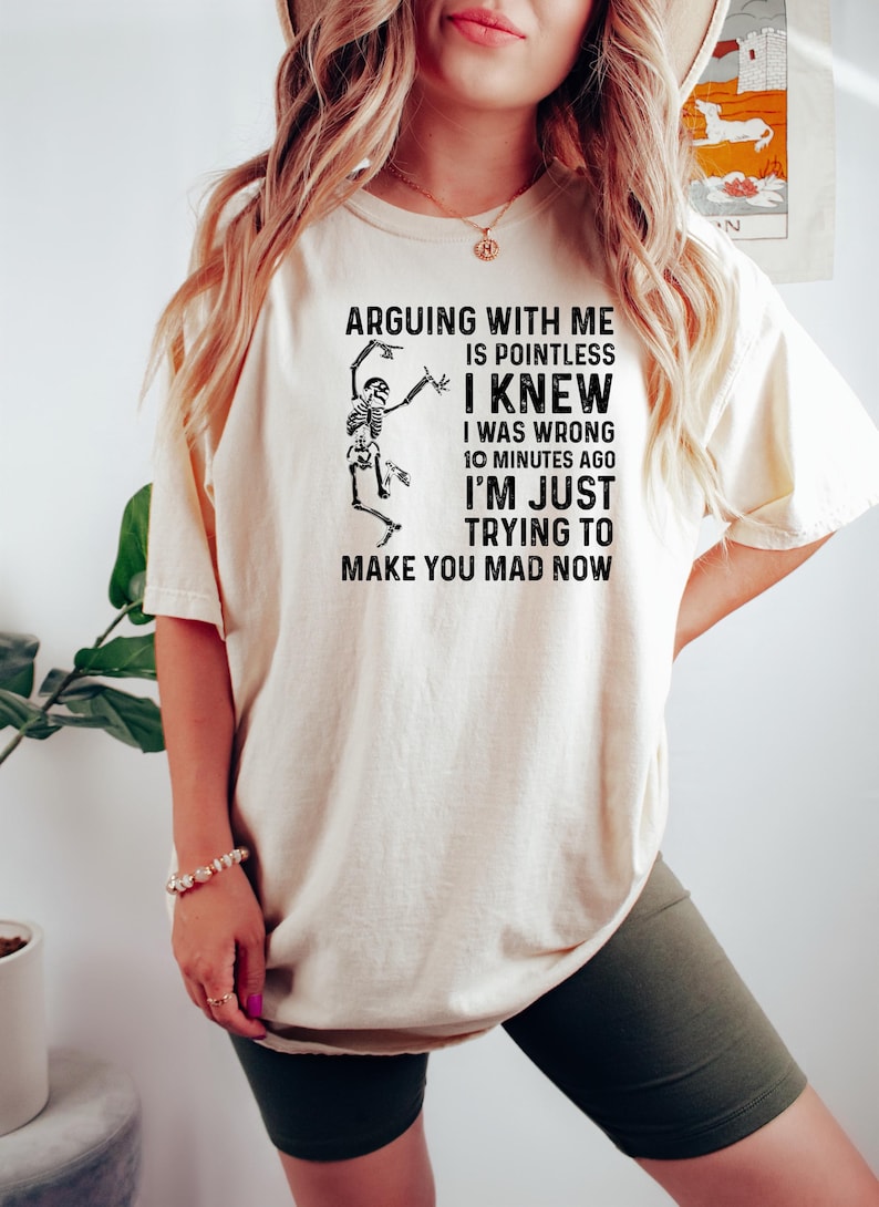 Puede incluir: Camiseta color crema con el texto "ARGUING WITH ME IS POINTLESS I KNEW I WAS WRONG 10 MINUTES AGO I'M JUST TRYING TO MAKE YOU MAD NOW" y un gr&aacute;fico de esqueleto. La camiseta se usa con pantalones cortos verde oscuro.