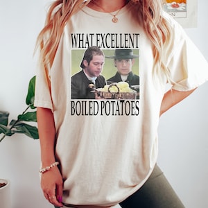 May include: A white t-shirt with the text "What excellent boiled potatoes" and a picture of two men in colonial clothing looking at a plate of potatoes.