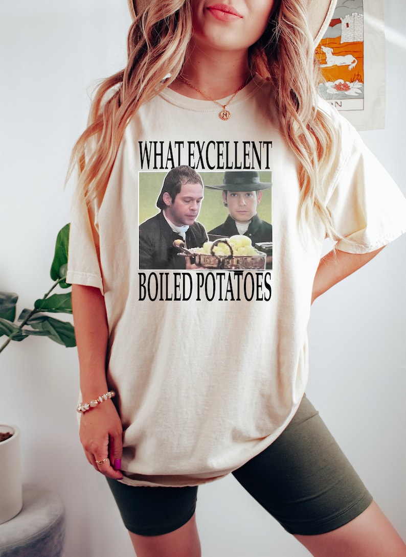 May include: A white t-shirt with the text "What excellent boiled potatoes" and a picture of two men in colonial clothing looking at a plate of potatoes.