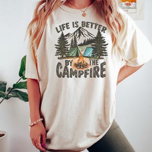 K&ouml;nnte beinhalten: Ein wei&szlig;es T-Shirt mit einem grafischen Aufdruck einer Bergszene mit einem Lagerfeuer, Zelten und B&auml;umen. Der Text "Life is better by the campfire" ist auf das Shirt gedruckt.