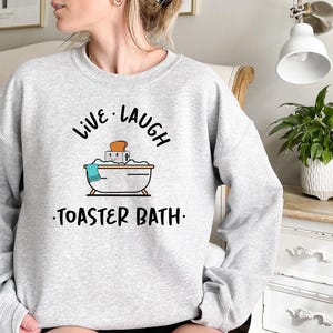 May include: Gray sweatshirt with the text "Live Laugh Toaster Bath" in black. The graphic features a cartoon bathtub with a slice of bread inside.