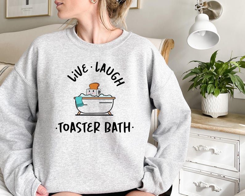 May include: Gray sweatshirt with the text "Live Laugh Toaster Bath" in black. The graphic features a cartoon bathtub with a slice of bread inside.
