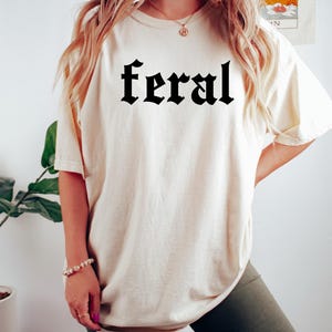 May include: A woman wearing a white oversized t-shirt with the word "feral" printed in black gothic lettering. She is also wearing dark green cycling shorts.