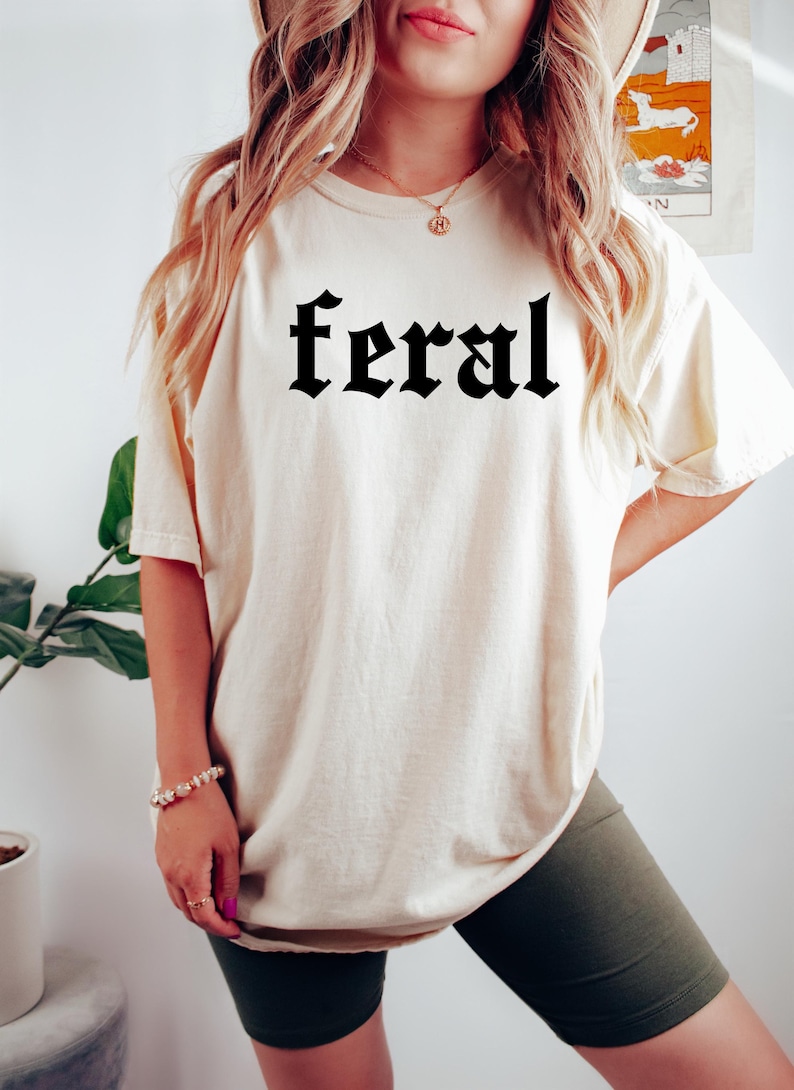 May include: A woman wearing a white oversized t-shirt with the word "feral" printed in black gothic lettering. She is also wearing dark green cycling shorts.