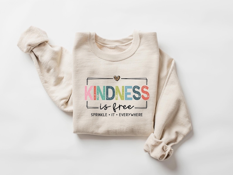 May include: A beige sweatshirt with the text "KINDNESS is free" in rainbow colors and a small leopard print heart. The text is framed in a black rectangle and the words "SPRINKLE IT EVERYWHERE" are printed below.