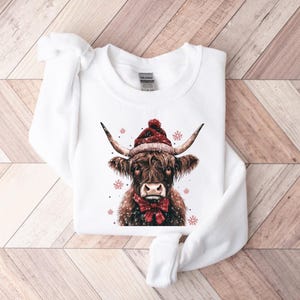 May include: White sweatshirt with a brown and white Highland cow wearing a red Santa hat. The cow is surrounded by snowflakes.