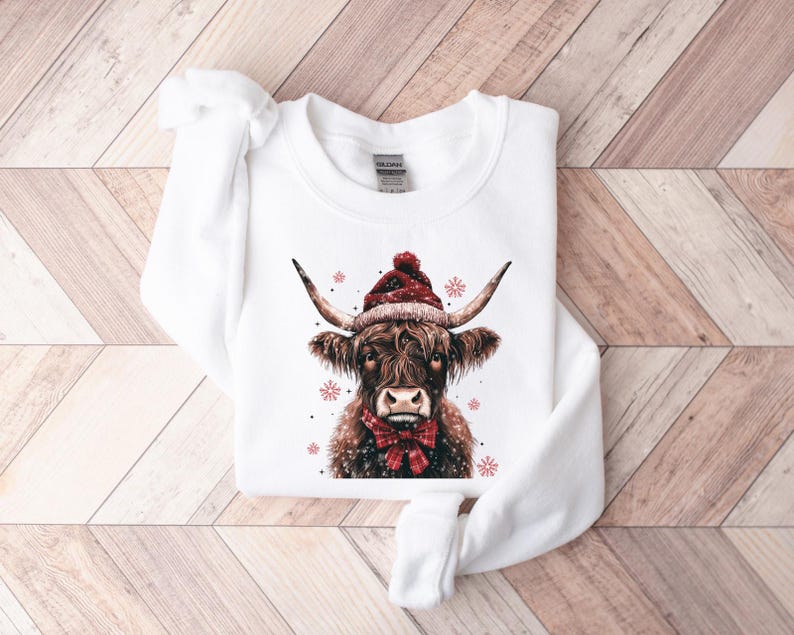 May include: White sweatshirt with a brown and white Highland cow wearing a red Santa hat. The cow is surrounded by snowflakes.