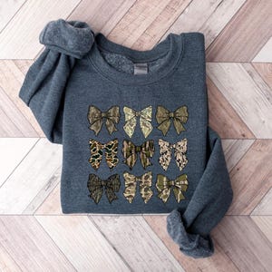 May include: A dark gray sweatshirt with a pattern of nine camouflage bows in shades of green, brown, and tan.