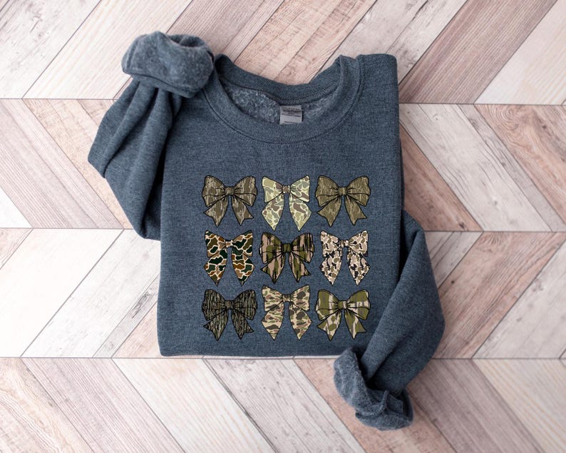May include: A dark gray sweatshirt with a pattern of nine camouflage bows in shades of green, brown, and tan.