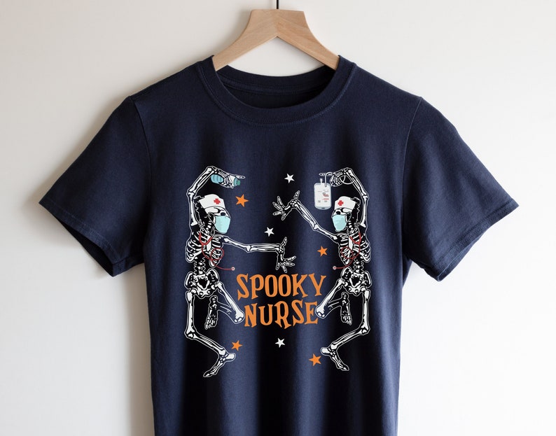 Puede incluir: Una camiseta azul marino con un gr&aacute;fico de dos esqueletos vestidos de enfermeras con mascarillas faciales y sosteniendo suministros m&eacute;dicos. El texto "Spooky Nurse" est&aacute; impreso en naranja en la camiseta.