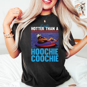 May include: Black t-shirt with the text "it gets HOTTER THAN A HOOCHIE COOCHIE" in white and blue. The graphic features a person in a cowboy hat floating on a purple and red inflatable tube in water.
