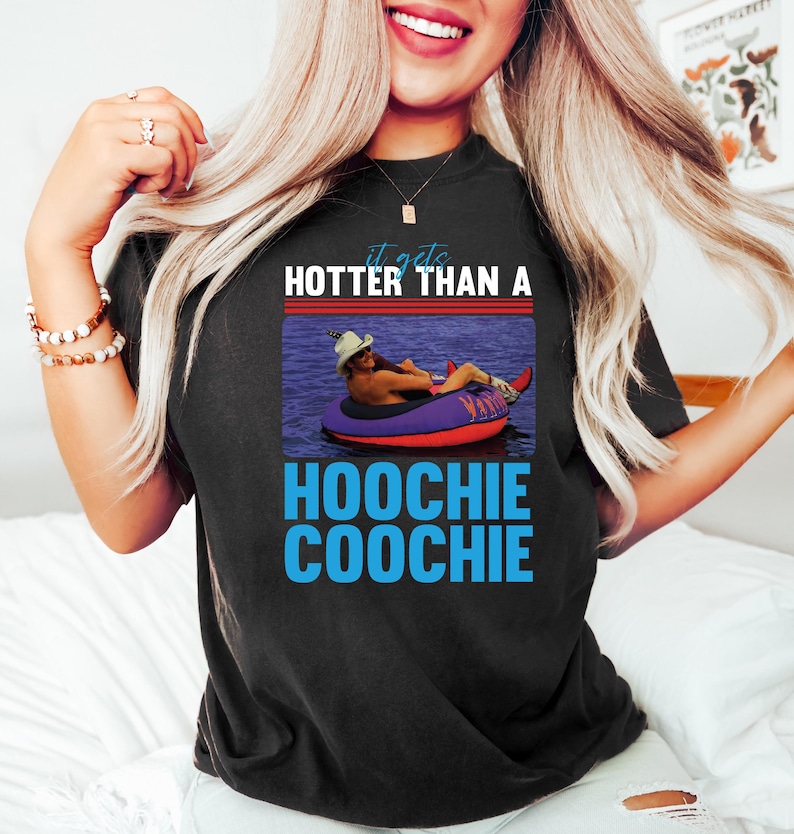 May include: Black t-shirt with the text "it gets HOTTER THAN A HOOCHIE COOCHIE" in white and blue. The graphic features a person in a cowboy hat floating on a purple and red inflatable tube in water.