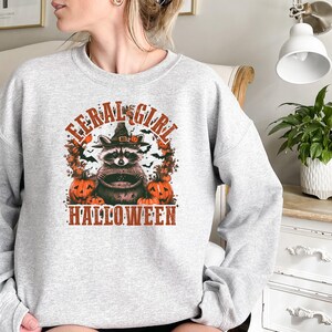 Pu&ograve; includere: Felpa grigia con una grafica di un procione che indossa un cappello da strega e tiene un calderone. Il procione &egrave; circondato da zucche e pipistrelli. Il testo "Feral Girl Halloween" &egrave; stampato sotto la grafica.