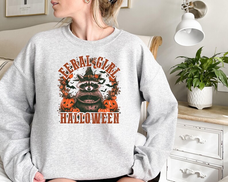Pu&ograve; includere: Felpa grigia con una grafica di un procione che indossa un cappello da strega e tiene un calderone. Il procione &egrave; circondato da zucche e pipistrelli. Il testo "Feral Girl Halloween" &egrave; stampato sotto la grafica.