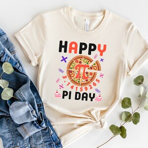 May include: A beige t-shirt with the text "Happy Pi Day" and a pizza slice with the mathematical symbol for pi in the center. The pizza slice is surrounded by numbers and math symbols.