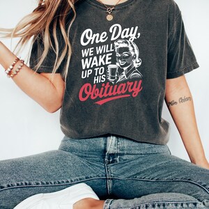 Puede incluir: Camiseta gris oscuro con el texto "One Day, We Will Wake Up To His Obituary" en blanco y rojo. La camiseta presenta una ilustraci&oacute;n de estilo vintage de una mujer sosteniendo una taza. La modelo lleva vaqueros azules y zapatos blancos.